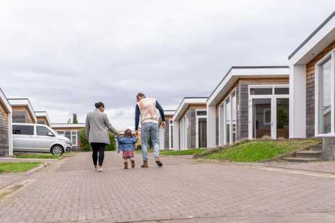 Een familie loopt samen met hun kind hand in hand op een pad tussen moderne lodges in een park.
