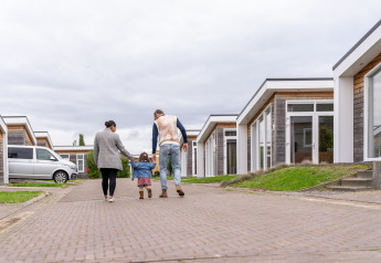 En familie går gennem et moderne hytteområde med en lille pige mellem sig på en overskyet dag.