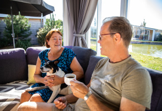 Dos personas se relajan con un perro en el sofá dentro de la Biebosch-lodge de Resort Mooi Bemelen, Países Bajos.