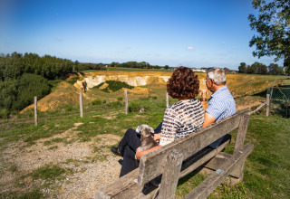 Et ældre par sidder på en bænk med deres hund og nyder udsigten ved Biebosch - tuinhuis på Resort Mooi Bemelen.