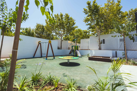 Playground with swings, slide, and carousel surrounded by trees at Camping Costa del Sol Glamping Village.