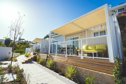Vista exterior de lodges modernos con terrazas de madera, sofás amarillos y plantas en un día soleado.