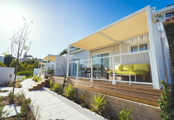 Vista exterior de lodges modernos con terrazas de madera, sofás amarillos y plantas en un día soleado.