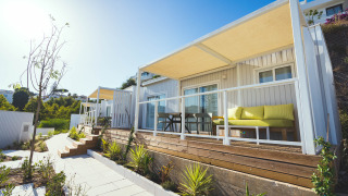 Vue extérieure de lodges modernes avec terrasse en bois, canapé jaune et plantes par une journée ensoleillée.