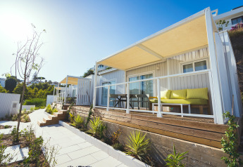 Udendørs billede af moderne lodges med træterrasse, gule sofaer og grøn beplantning en solrig dag.