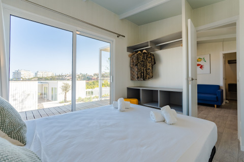 Chambre dans un lodge avec lit double, armoire ouverte, canapé bleu et vue sur des bâtiments modernes.