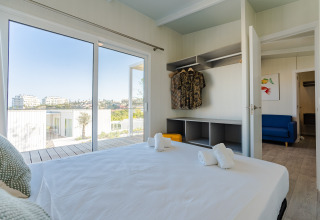 Dormitorio en una cabaña con cama doble, armario abierto, sofá azul y vistas a edificios modernos.