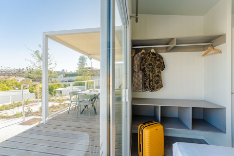 Udsigt fra et lodgeværelse med åben garderobe, gult kuffert og terrasse med stole og bord udenfor.