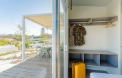 Udsigt fra et lodgeværelse med åben garderobe, gult kuffert og terrasse med stole og bord udenfor.