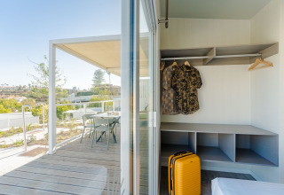 Zicht vanuit een lodgekamer met open kast, hemden, gele koffer en terras met stoelen buiten.