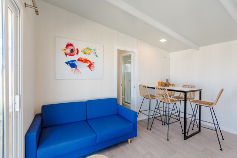 Licht lodge-interieur met blauwe sofa, kleurrijk viskunstwerk en hoge tafel met vier gevlochten stoelen.