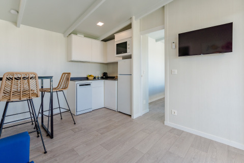 Modern lodge interior with compact kitchen, high chairs, wall-mounted TV and light wooden floors.