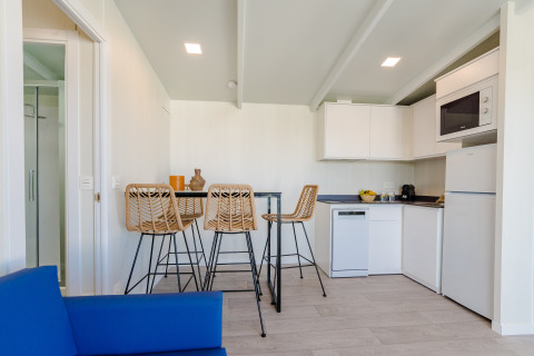 Cocina moderna en lodge con cuatro taburetes de mimbre, mesa alta, muebles blancos y sofá azul al frente.