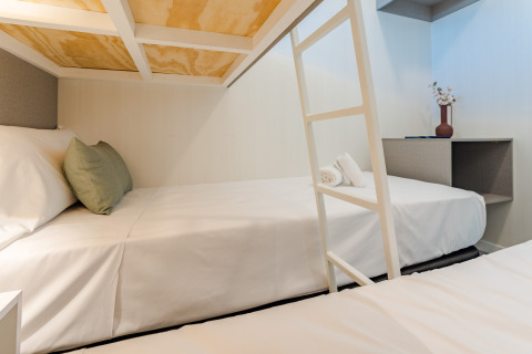 Habitación moderna de albergue con literas, sábanas blancas, cojín verde y decoración minimalista con florero.