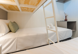 Habitación moderna de albergue con literas, sábanas blancas, cojín verde y decoración minimalista con florero.