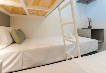 Habitación moderna de albergue con literas, sábanas blancas, cojín verde y decoración minimalista con florero.