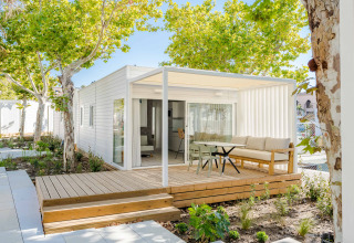 Modern lodge with a wooden deck, outdoor seating area, and lush green trees providing shade.