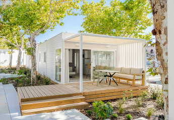Modern lodge with a wooden deck, outdoor seating area, and lush green trees providing shade.