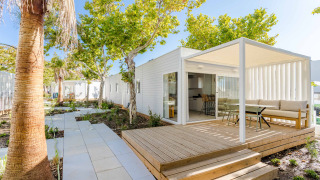 Lodge moderne avec terrasse couverte, coin salon extérieur et arbres verdoyants sous un ciel bleu vif.