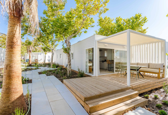 Moderne lodge med overdækket terrasse, udendørs siddepladser og grønne træer under klar blå himmel.
