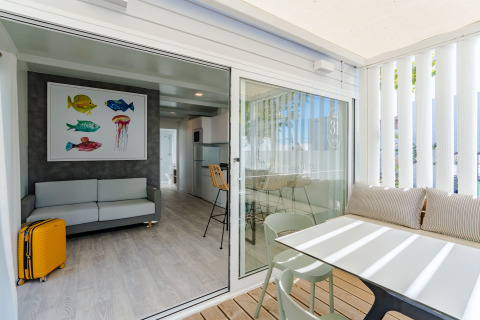 Bright lodge room featuring a sofa, colorful fish artwork, yellow suitcase, and terrace with dining area.