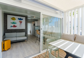 Bright lodge room featuring a sofa, colorful fish artwork, yellow suitcase, and terrace with dining area.
