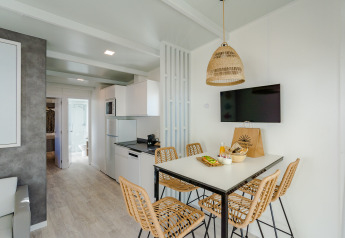 Modern lodge living space with dining table, wicker chairs, a kitchen area, wall-mounted TV, and natural light.