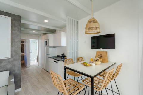 Modern lodge living space with dining table, wicker chairs, a kitchen area, wall-mounted TV, and natural light.