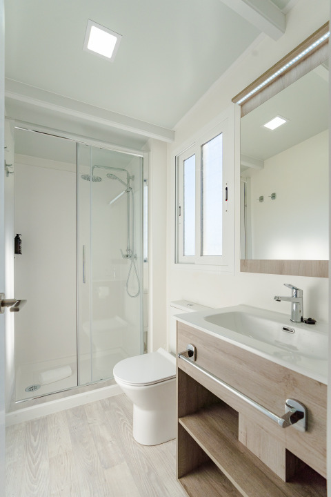 Baño moderno en una cabaña con ducha de vidrio, inodoro, lavabo, espejo grande y ventana luminosa.