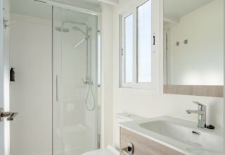 Baño moderno en una cabaña con ducha de vidrio, inodoro, lavabo, espejo grande y ventana luminosa.