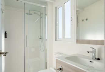 Baño moderno en una cabaña con ducha de vidrio, inodoro, lavabo, espejo grande y ventana luminosa.