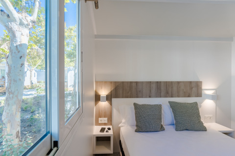 Chambre lumineuse en lodge avec grand lit, coussins verts et large fenêtre donnant sur des arbres et la nature.