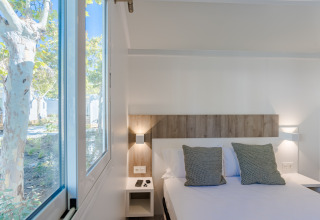 Chambre lumineuse en lodge avec grand lit, coussins verts et large fenêtre donnant sur des arbres et la nature.