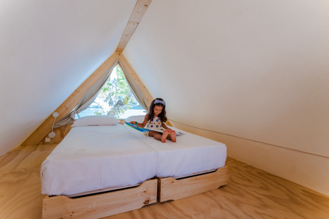 Una bambina legge un libro su un letto in una lodge a forma di A al Camping Costa del Sol Glamping Village, Spagna.