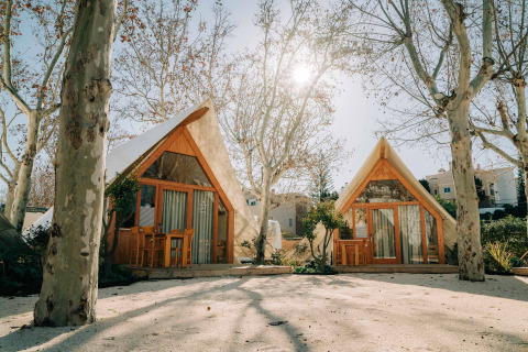 Zwei A-förmige Holzhütten unter kahlen Bäumen im Sonnenlicht im Camping Costa del Sol Glamping Village.