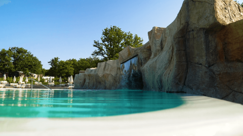 Piscina al Camping Lanterna in Istria, Croazia, con parete rocciosa, cascata e lettini sotto il cielo azzurro.