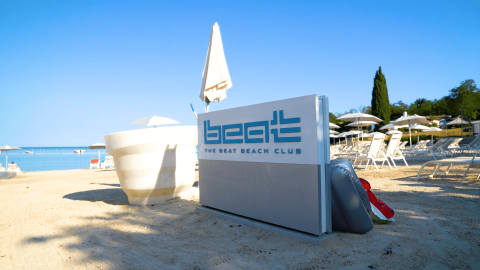 Beach club sign and sun loungers on sandy shore at Camping Lanterna, Istria, Croatia, under a clear blue sky.