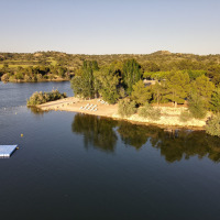 Zona de lago con playa de arena y tumbonas cerca de Caspe, Aragón, España, rodeada de árboles y colinas.