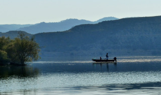 Personas pescando desde una pequeña barca en un lago tranquilo cerca de Caspe, Aragón, España, con colinas al fondo.