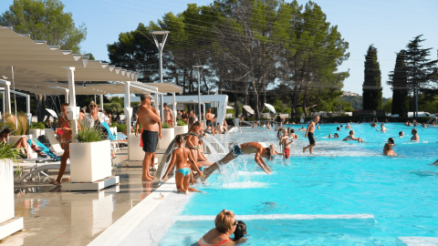 Feriepark poolområde med folk, børn der springer i vandet, parasoller og liggestole ved Camping Valkanela.