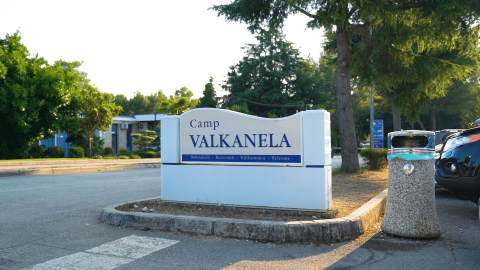 Toegangsbord van Camp Valkanela, een vakantiepark in Istrië, Kroatië, omgeven door bomen en geparkeerde auto’s.