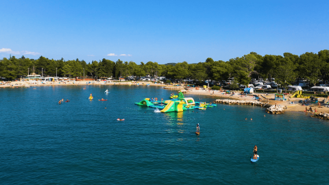 Spiaggia e parco acquatico galleggiante al Camping Valkanela, Istria, Croazia, con persone e pini sullo sfondo.