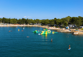 Spiaggia e parco acquatico galleggiante al Camping Valkanela, Istria, Croazia, con persone e pini sullo sfondo.