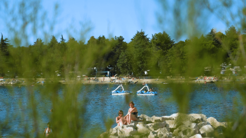 Vista della spiaggia di Camping Valkanela in Istria, Croazia, con persone e pedalò sul lago tra il verde.