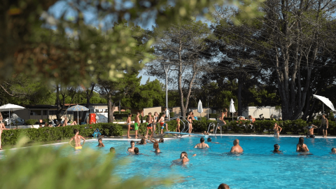 Personas disfrutan de un día soleado en la piscina de Camping Valkanela, rodeada de árboles y cabañas.