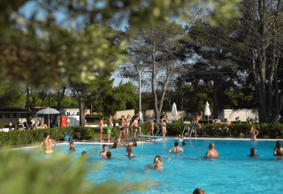 Personas disfrutan de un día soleado en la piscina de Camping Valkanela, rodeada de árboles y cabañas.