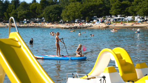 Niños jugando sobre una tabla de paddle surf en Camping Valkanela, Istria, Croacia, con la playa y gente al fondo.
