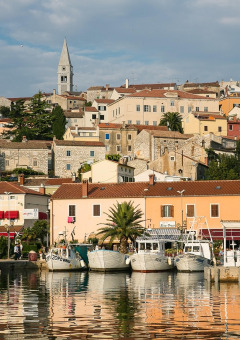 Foto de un pintoresco pueblo costero con barcos, casas coloridas, árboles y una torre de iglesia al fondo.