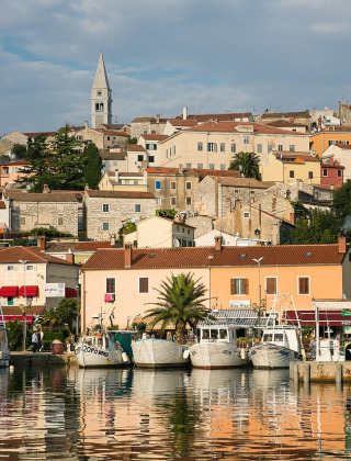 Foto van een sfeervol kustplaatsje met boten, kleurrijke huizen, groen en een kerktoren op de achtergrond.