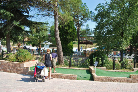 Familia jugando minigolf en Altomincio Family Park, Lombardía, Italia, con piscina y árboles al fondo.